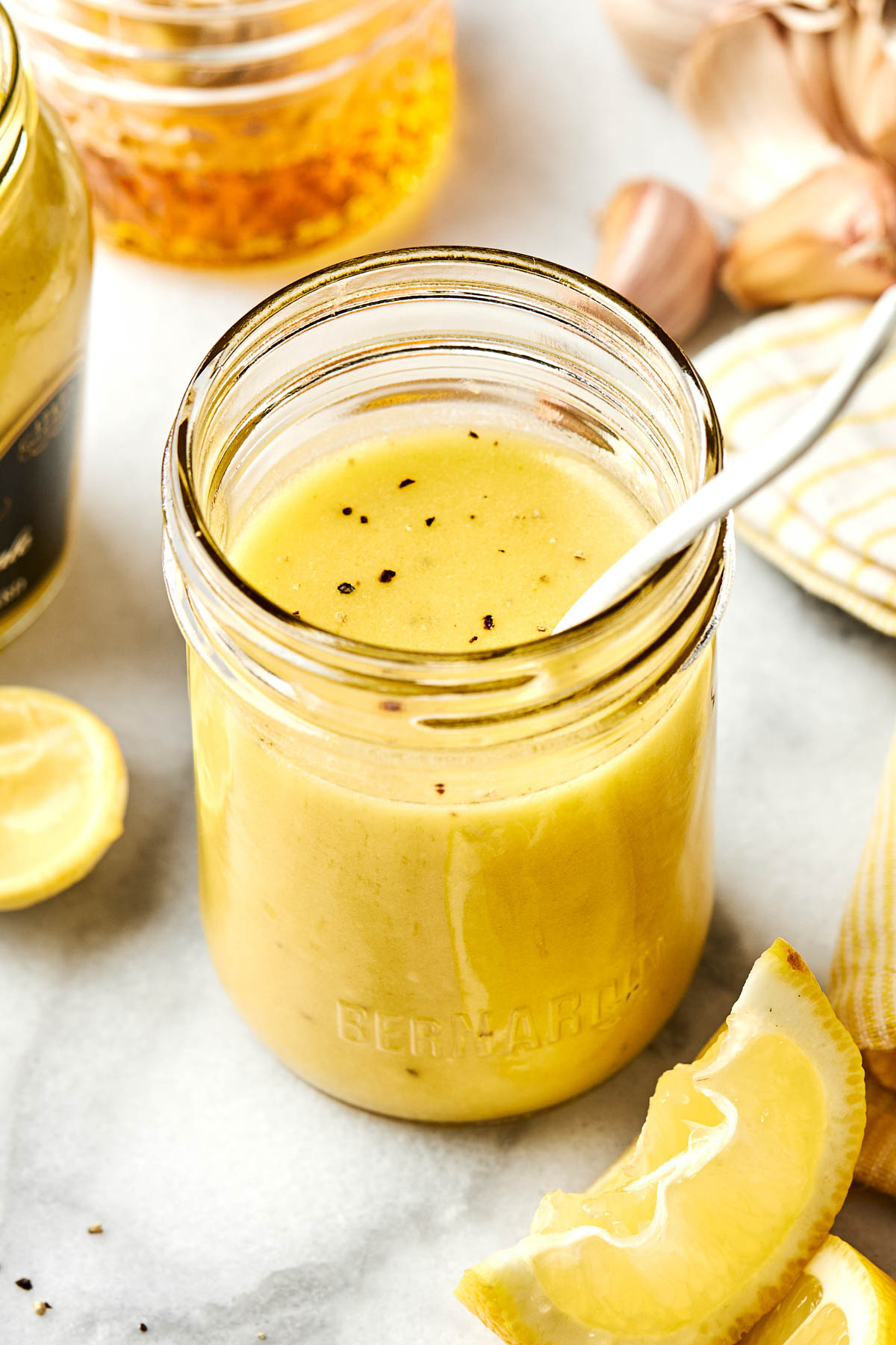 A glass jar filled with yellow salad dressing sits on a white surface, with a spoon inside. Lemon wedges, garlic cloves, and another jar are nearby. Black pepper is sprinkled on top of the dressing.