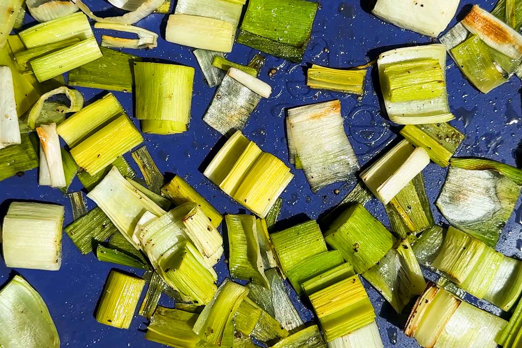 Chopped green and white pieces of leek scattered on a blue surface, with some pieces glistening as if freshly washed or lightly oiled.