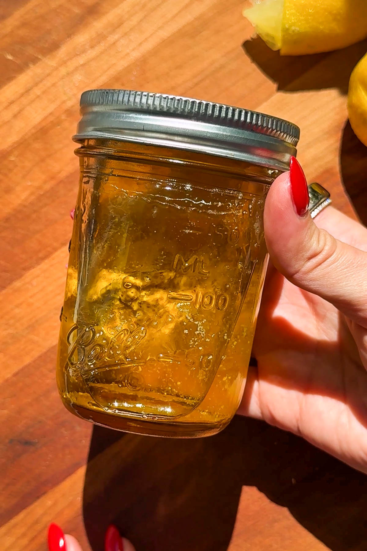 A hand with red nails holds a small glass jar of golden yellow jelly or honey, with sunlight reflecting off the jar and a wooden surface and slices of lemon in the background.