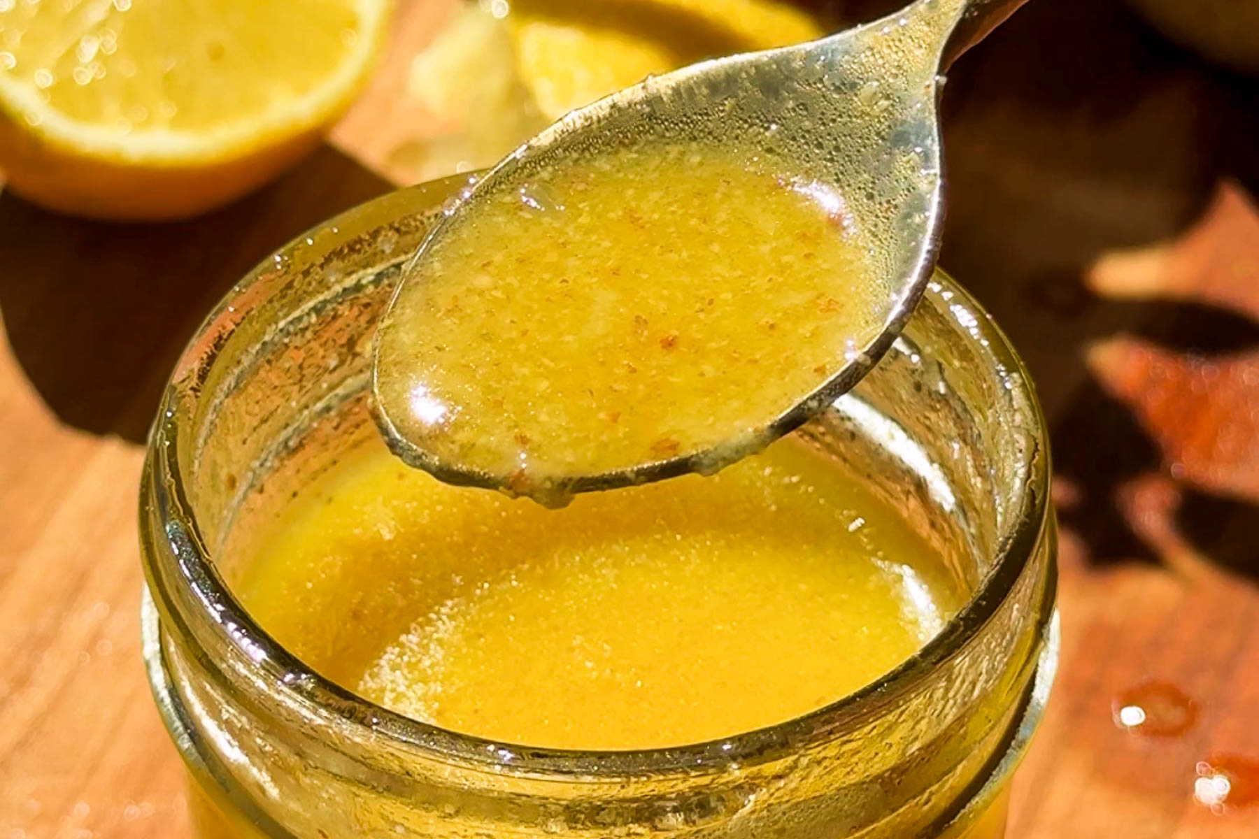 A close-up of a jar filled with yellow vinaigrette, with a spoon holding some of the dressing above the jar. Sliced lemon is visible in the background on a wooden surface.