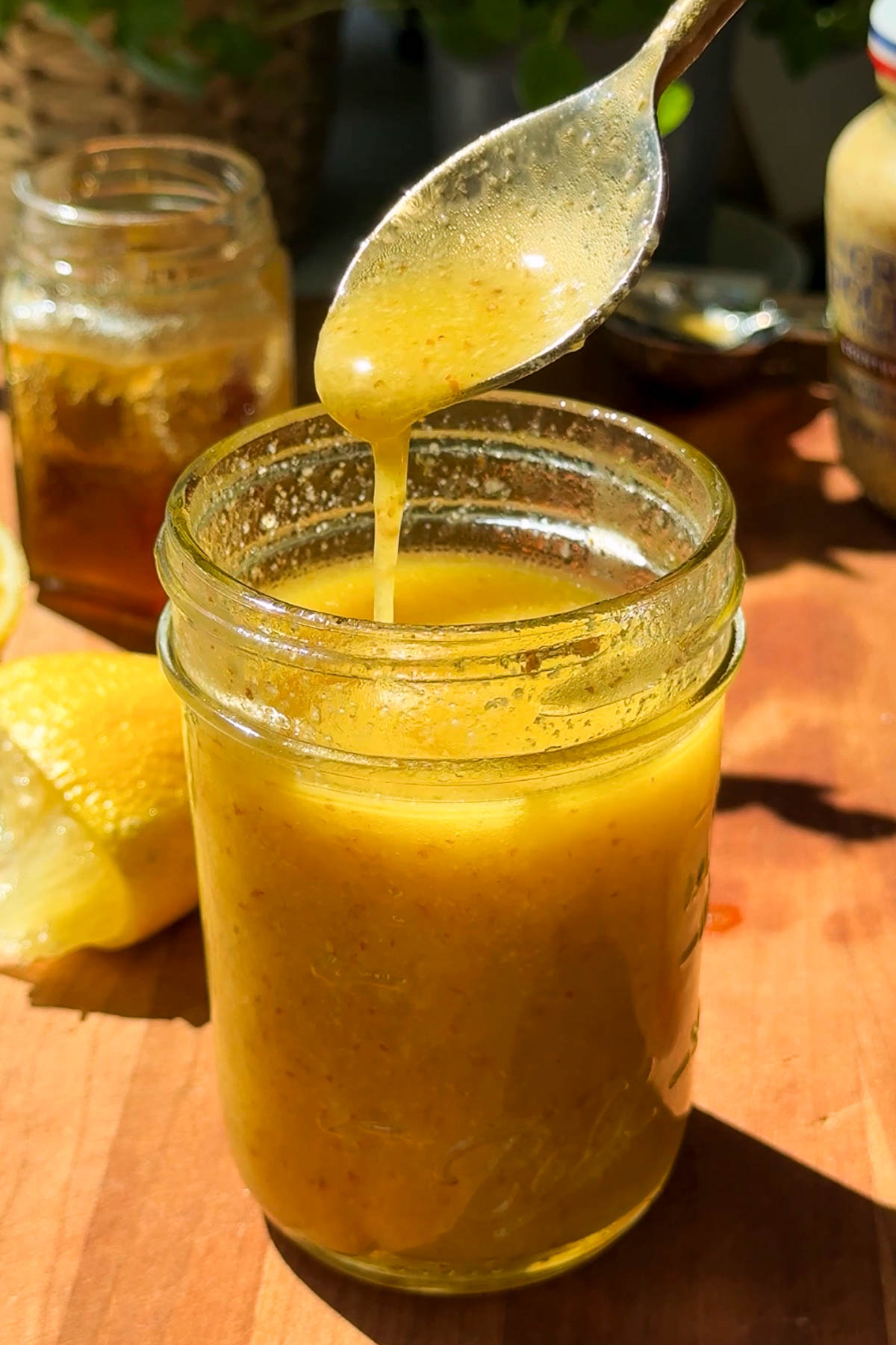 A spoon drizzles golden honey mustard dressing into a glass jar, with a halved lemon and jars of honey and mustard nearby on a sunlit wooden surface.