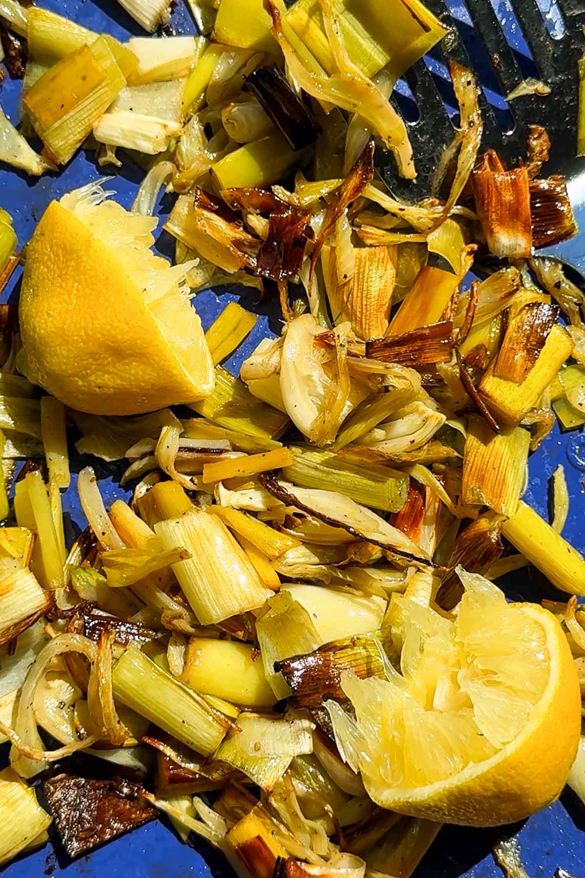 Close-up of roasted chopped leeks and garlic on a blue surface, with two squeezed lemon halves among the vegetables. Some pieces are caramelized and darkened, indicating thorough roasting.