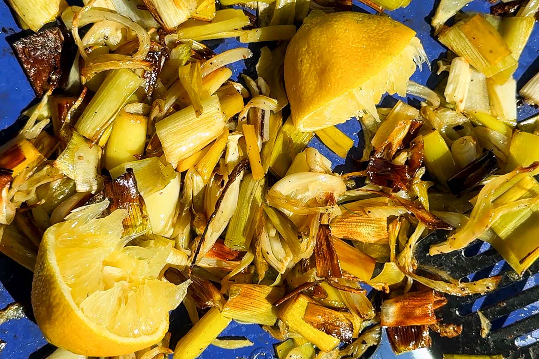Chopped and roasted leeks with charred edges and two squeezed lemon halves on a blue surface, glistening with oil and seasoning in bright sunlight.