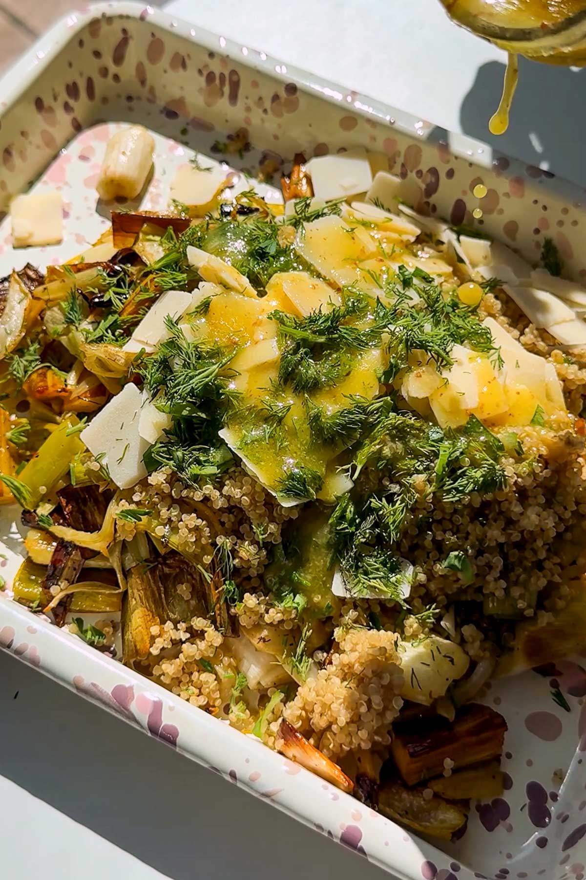 A close-up of a colorful grain salad with quinoa, roasted vegetables, shaved cheese, fresh herbs, and a drizzle of yellow dressing, served on a patterned plate.