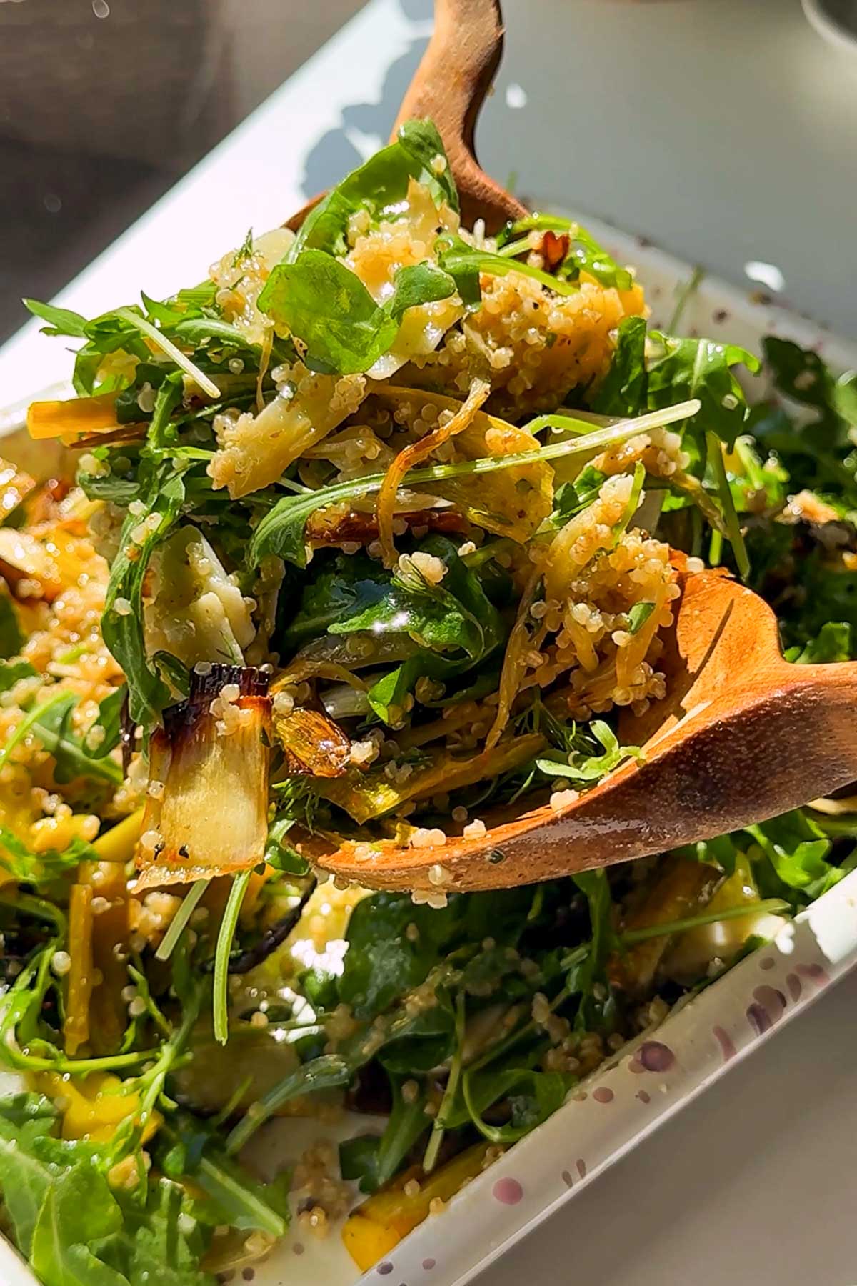 A wooden spoon serves a fresh salad with quinoa, leafy greens, grilled vegetables, and herbs on a white rectangular plate, illuminated by natural sunlight.