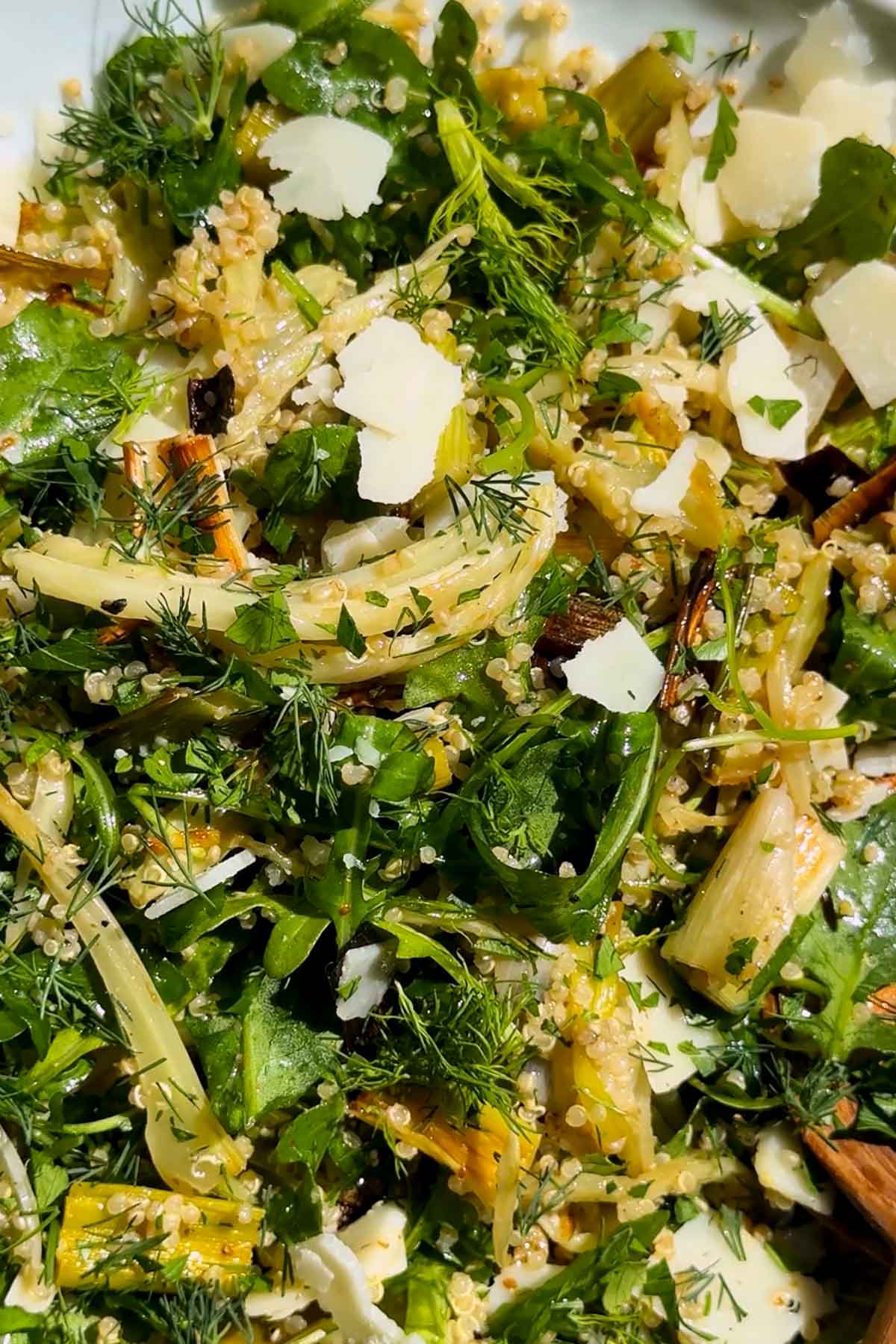 A close-up of a fresh salad with leafy greens, roasted vegetables, shaved parmesan cheese, quinoa, and herbs, all tossed together and garnished with dill.