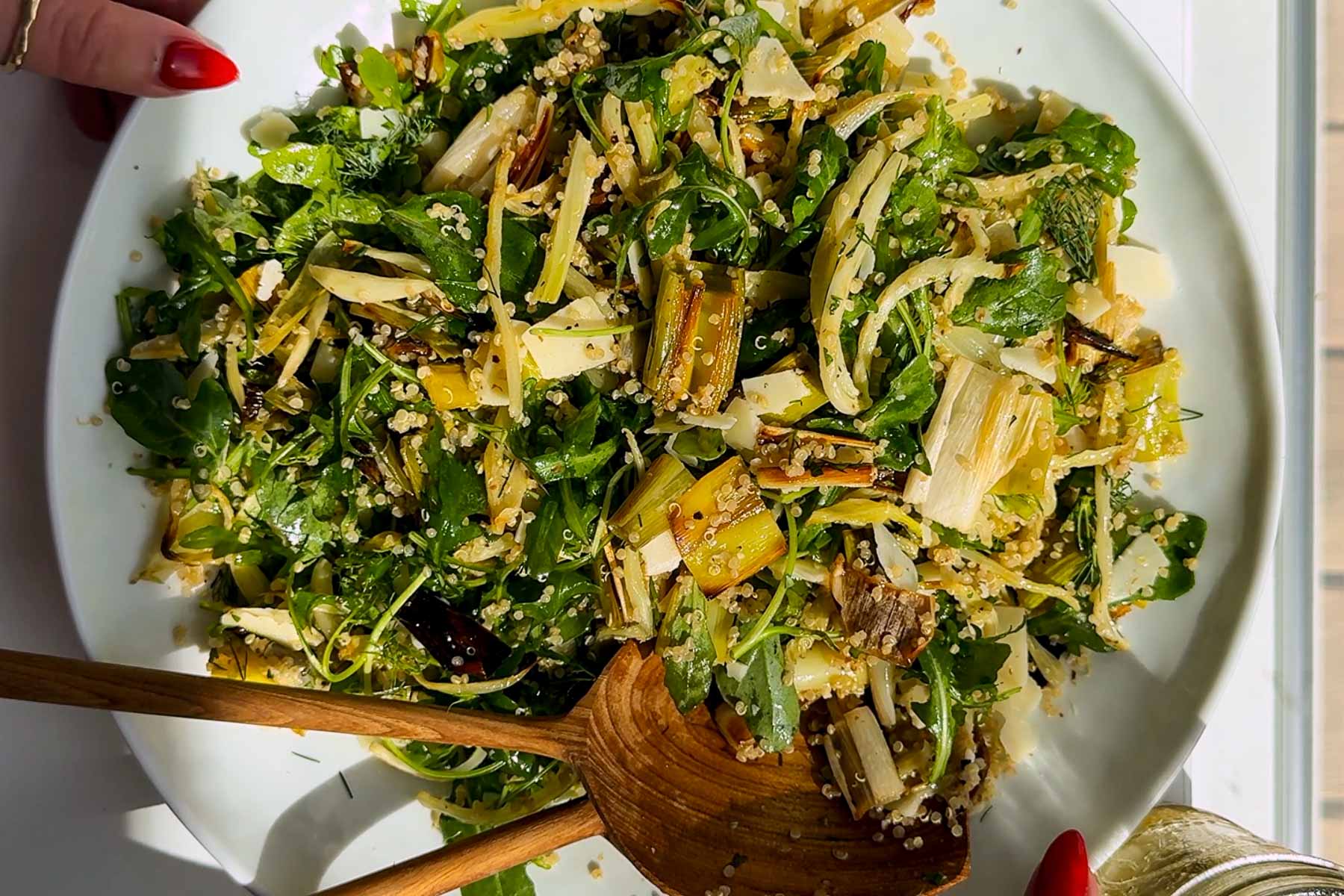 A plate of fresh salad with leafy greens, quinoa, thinly sliced vegetables, and cheese, being served with a wooden spoon. A hand with red nails is holding the plate’s edge.