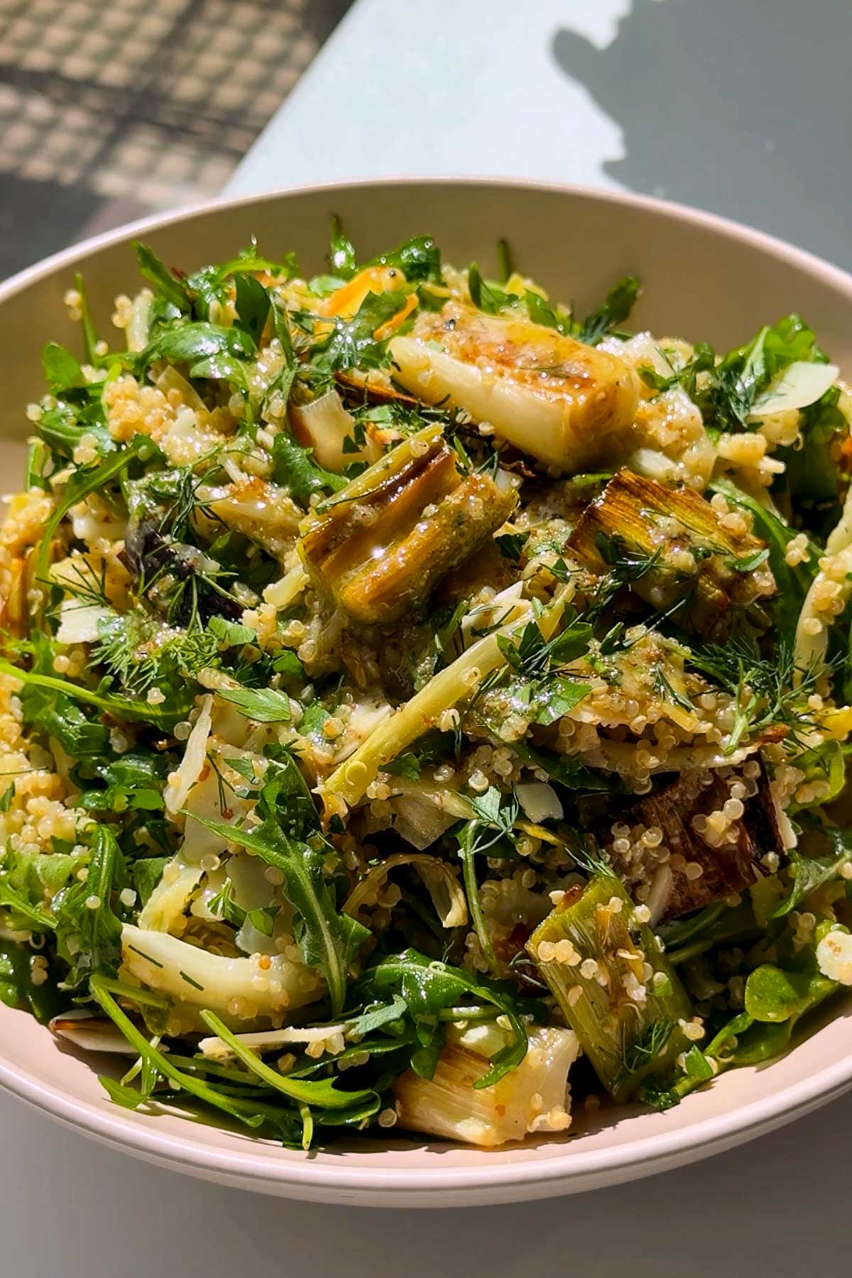 A bowl of fresh salad with quinoa, arugula, grilled leeks, and herbs, tossed in a light dressing, served in natural sunlight.