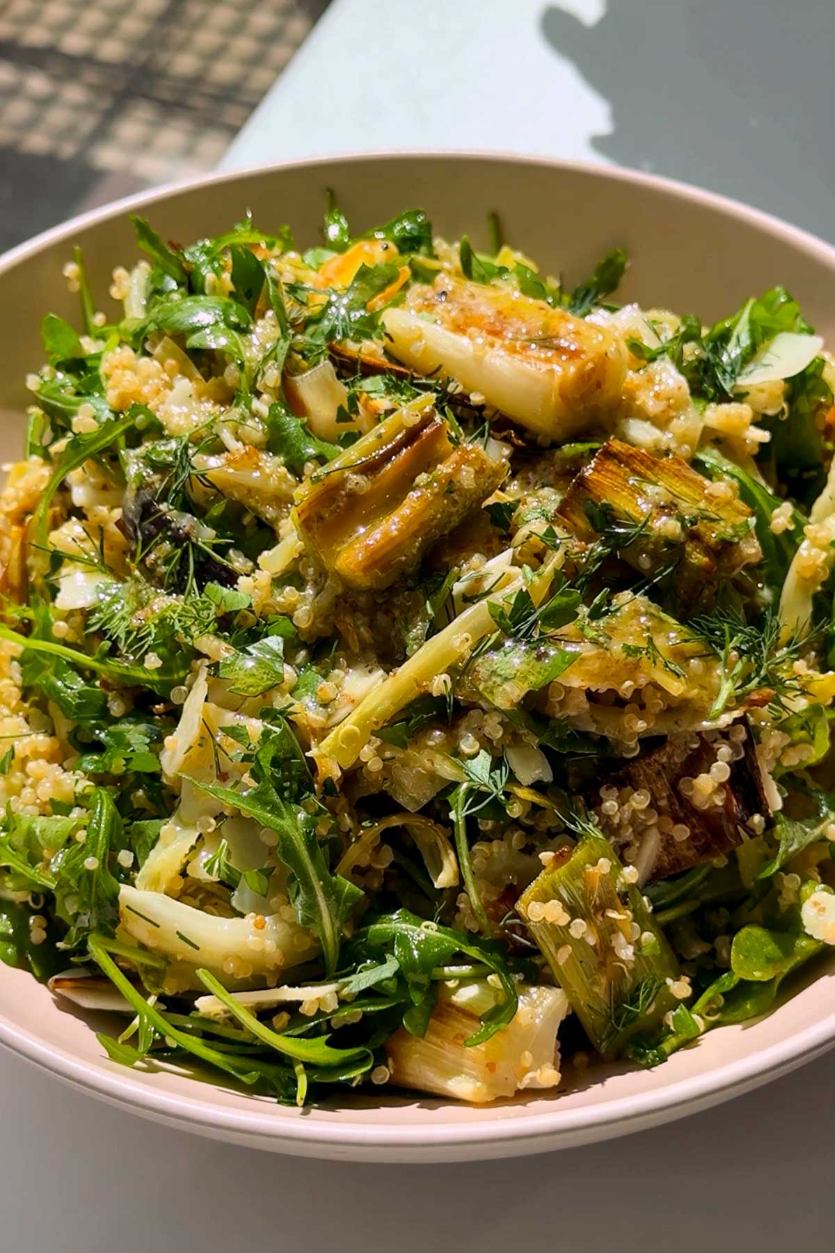 A bowl of fresh salad with leafy greens, quinoa, grilled vegetables, and herbs, served in natural sunlight.