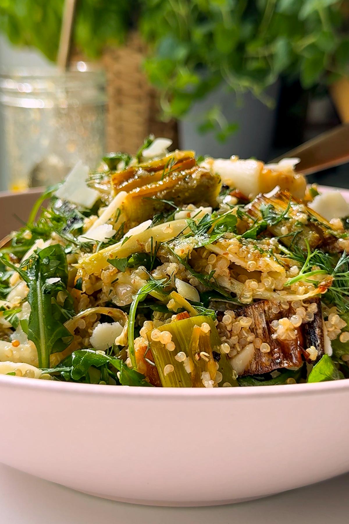 A close-up of a fresh quinoa salad in a white bowl, featuring arugula, shaved parmesan, roasted vegetables, and herbs, with a blurred green plant in the background.