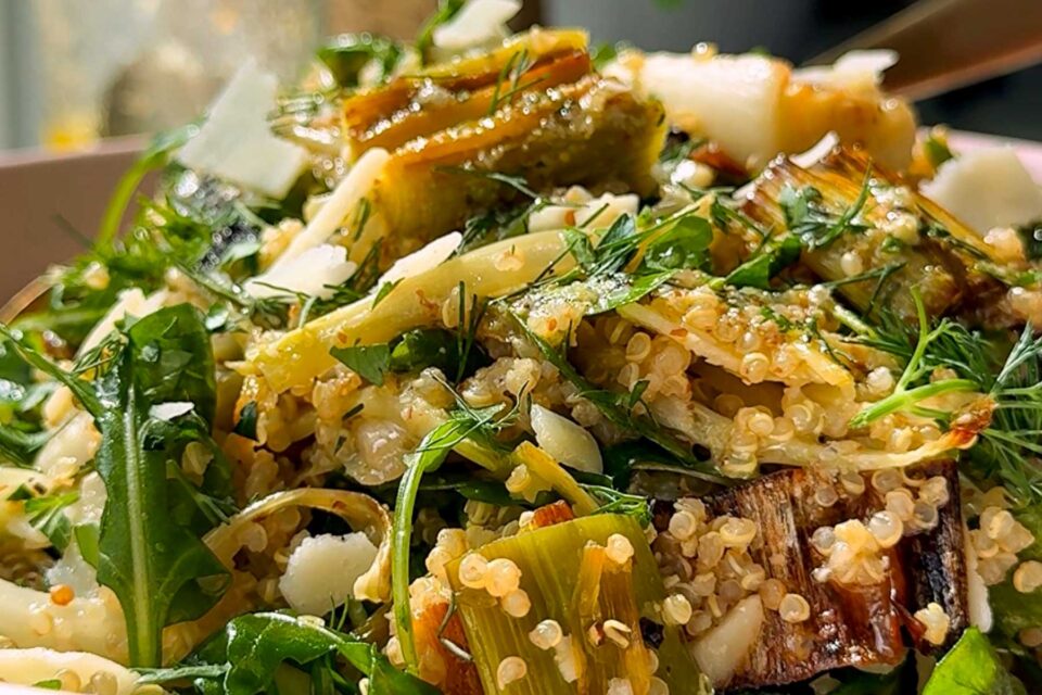 Close-up of a fresh quinoa salad with arugula, grilled vegetables, fresh herbs, and shaved parmesan cheese, showcasing vibrant green leaves, grains, and browned veggie pieces.