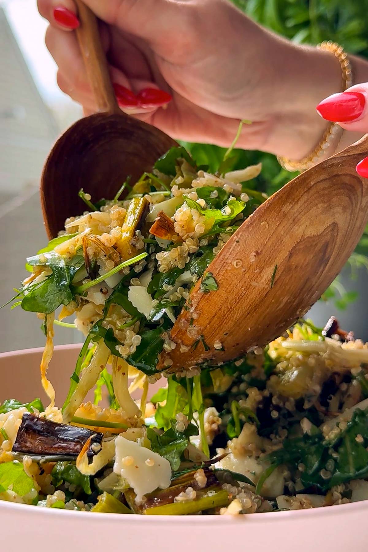 A close-up of a person with red nails serving a fresh quinoa salad with greens and grilled vegetables from a wooden bowl using wooden utensils.