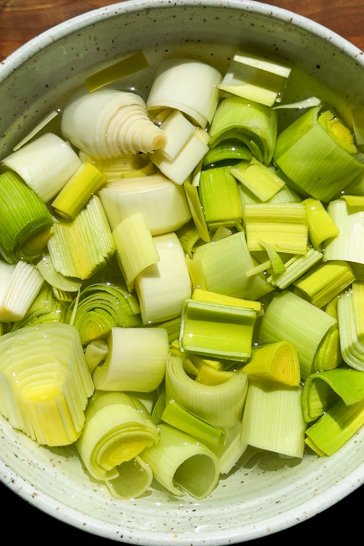 A bowl filled with chopped leeks soaking in water, showing green and white rings of the vegetable with sunlight highlighting their fresh, layered texture.