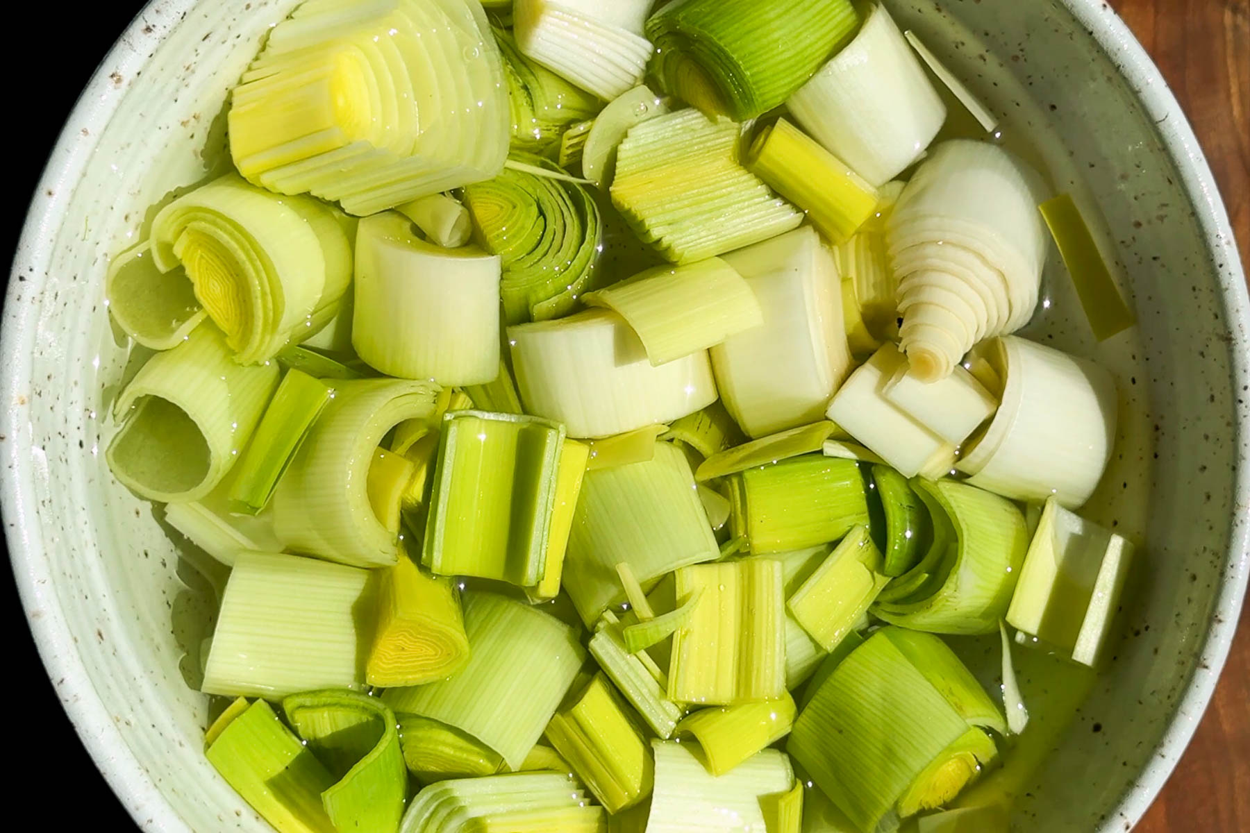 A bowl filled with chopped leeks soaking in water. The leeks are cut into round and half-moon pieces, displaying shades of green and white.