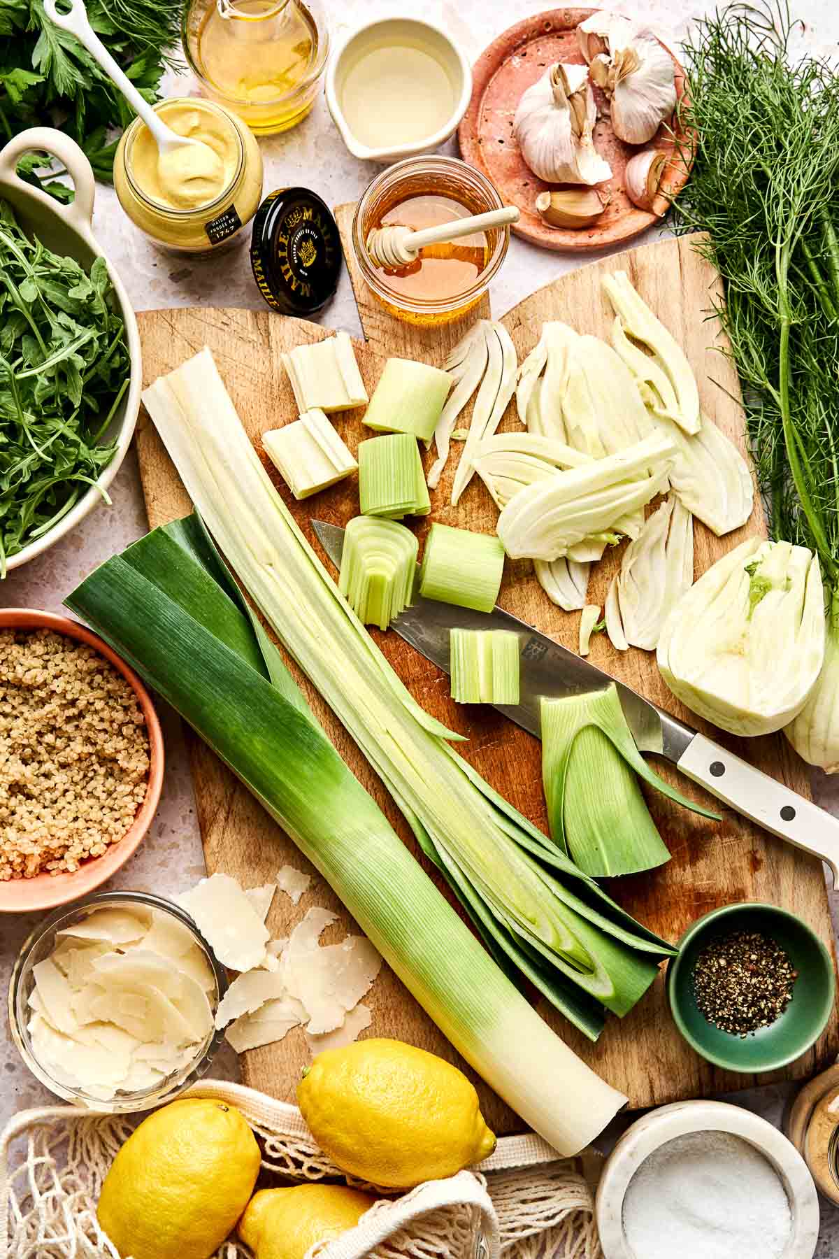 Overhead view of a wooden cutting board with sliced leek and fennel, surrounded by arugula, whole and sliced lemons, quinoa, Parmesan, Dijon mustard, honey, garlic, fresh dill, salt, and pepper.