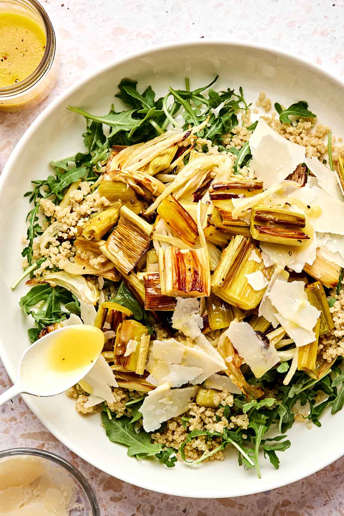 A white bowl filled with arugula, quinoa, roasted leeks, and shaved parmesan cheese, with a spoon drizzling dressing over the salad. A jar of dressing is nearby on the table.