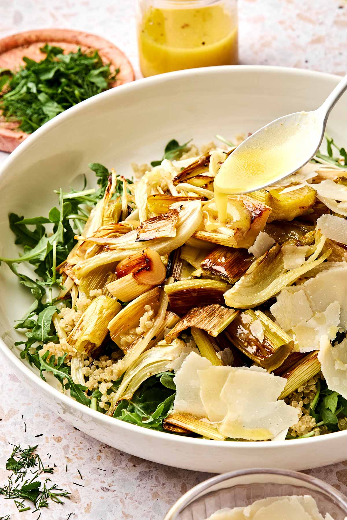 A white bowl of salad with roasted leeks and fennel, quinoa, arugula, and shaved parmesan. A spoon drizzles lemon dijon vinaigrette on top. Fresh herbs and a jar of dressing are in the background.