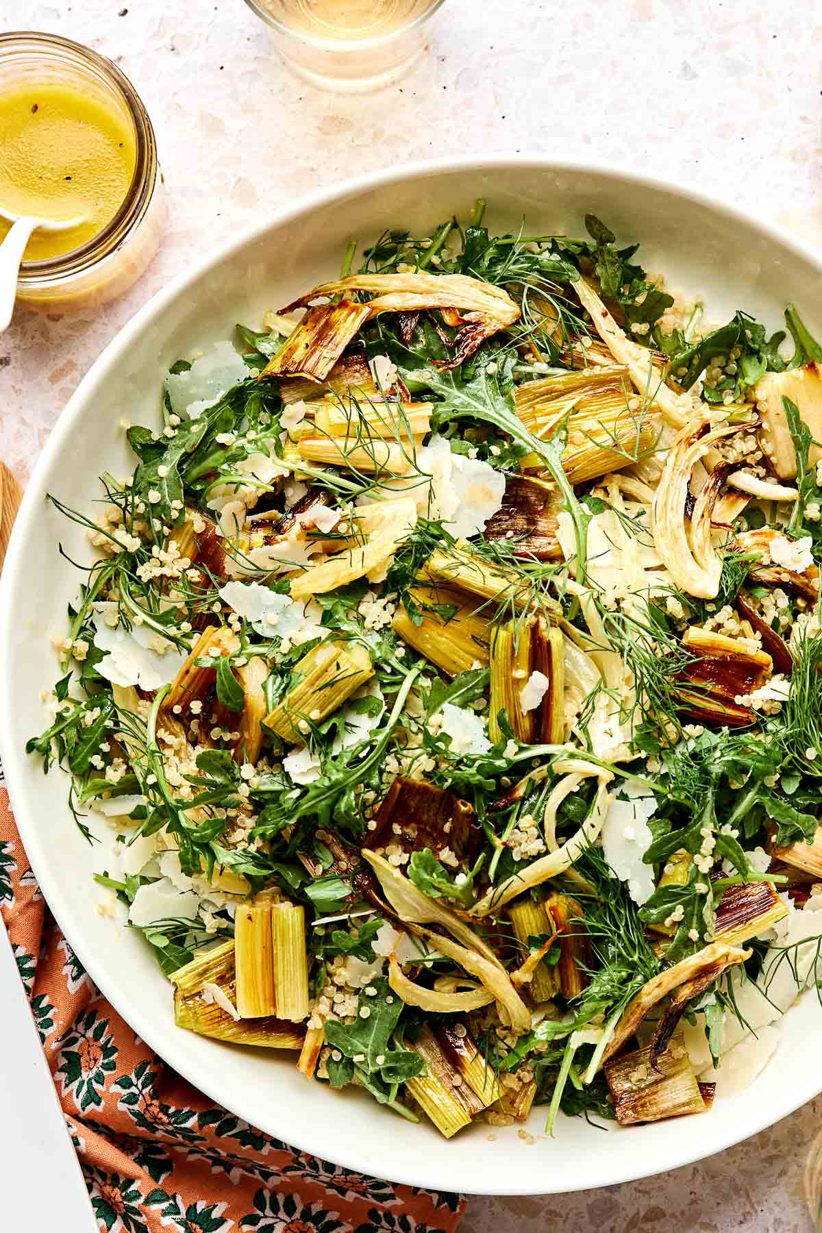 A bowl of leek salad with arugula, roasted vegetables, shaved Parmesan cheese, fresh dill, and grains, placed on a patterned napkin with a glass of dressing and a drink nearby.