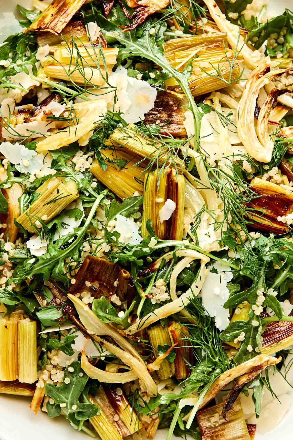 A close-up of a salad with roasted leeks, leafy greens, fresh dill, quinoa, and shaved parmesan cheese. The ingredients are mixed together, creating a vibrant and fresh appearance.