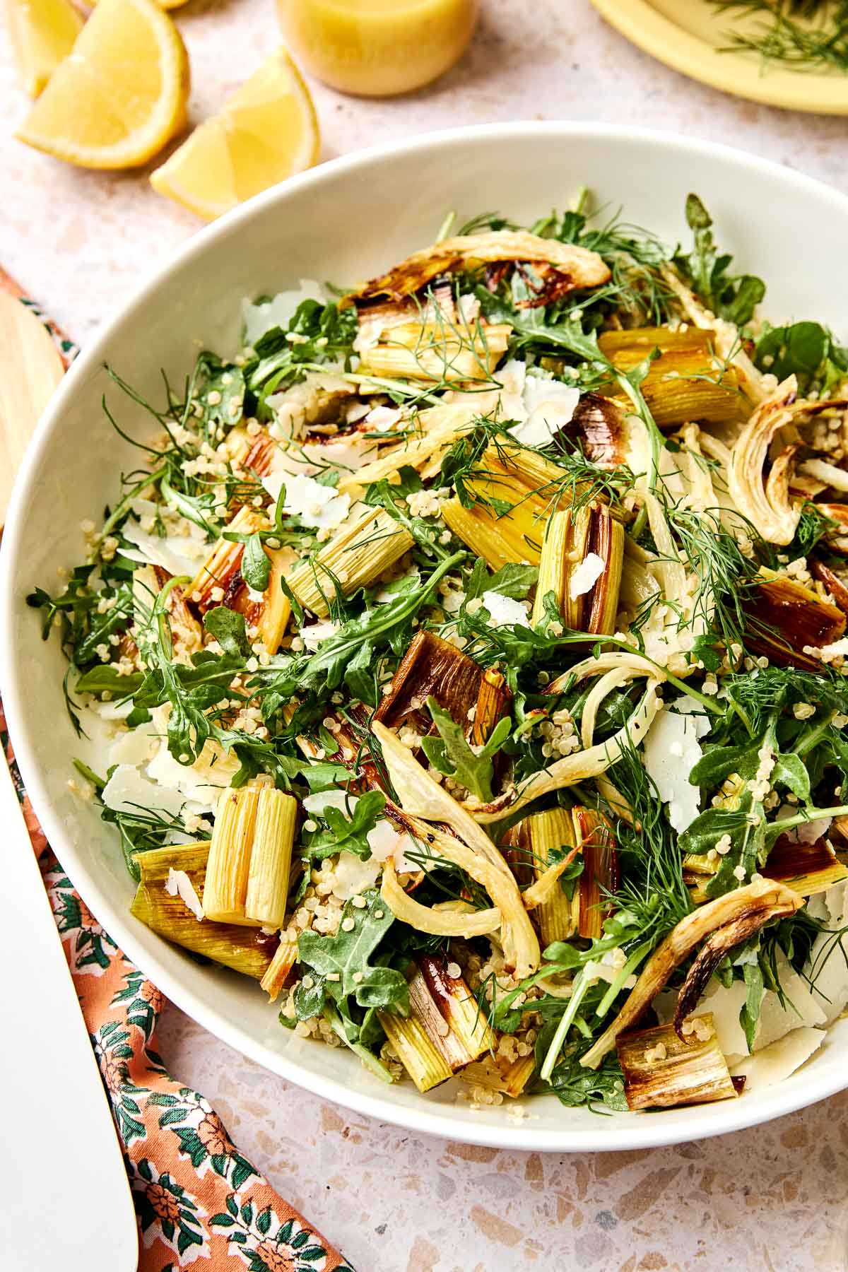A bowl of roasted leek salad featuring fennel, arugula, fresh herbs, and grated cheese, with lemon wedges and a floral napkin nearby.