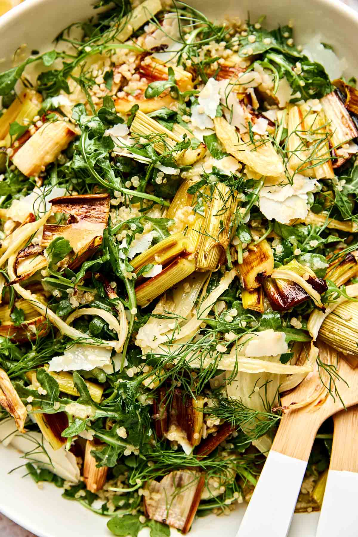 A close-up of a leeks salad with arugula, shaved parmesan, quinoa, and fresh herbs in a white bowl with wooden salad servers.