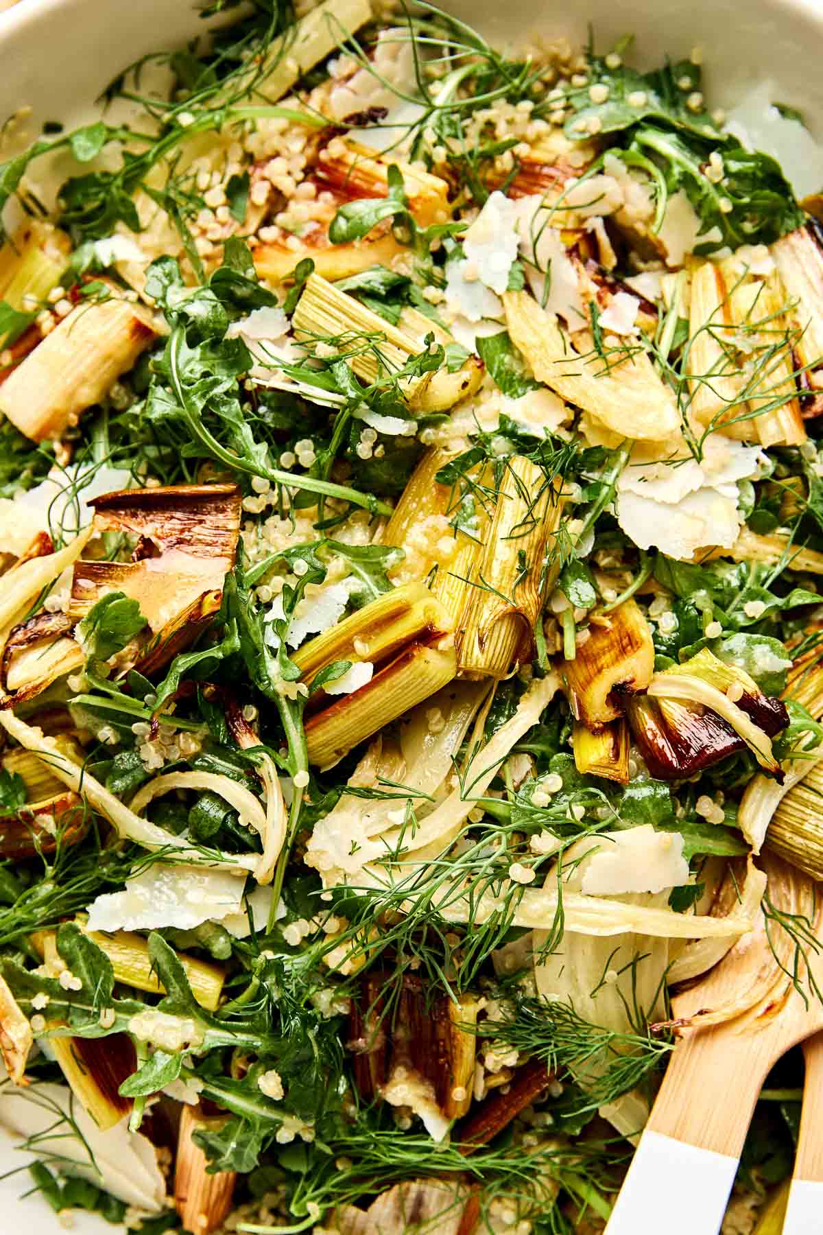 A close-up of a fresh salad with roasted leeks, arugula, shaved parmesan, fresh dill, and cooked quinoa, served in a white bowl with a wooden serving spoon.