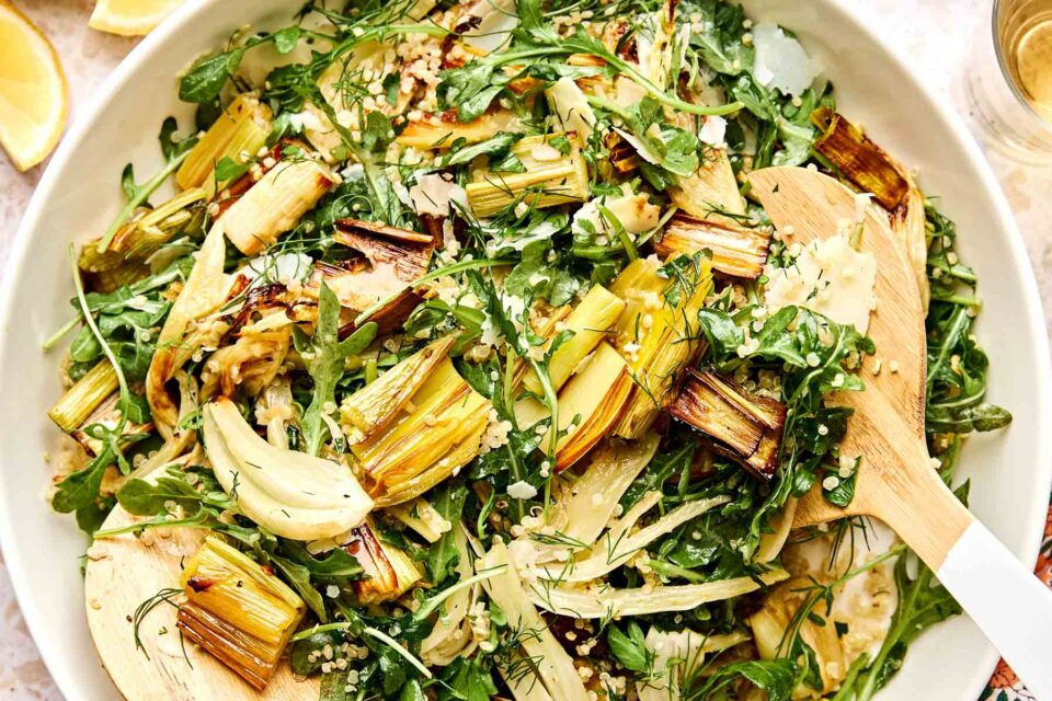 A close-up of a fennel and leek salad with arugula, herbs, and quinoa, tossed in a white bowl. A wooden spoon and spatula are mixing the ingredients. Lemon wedges are visible at the edge of the dish.