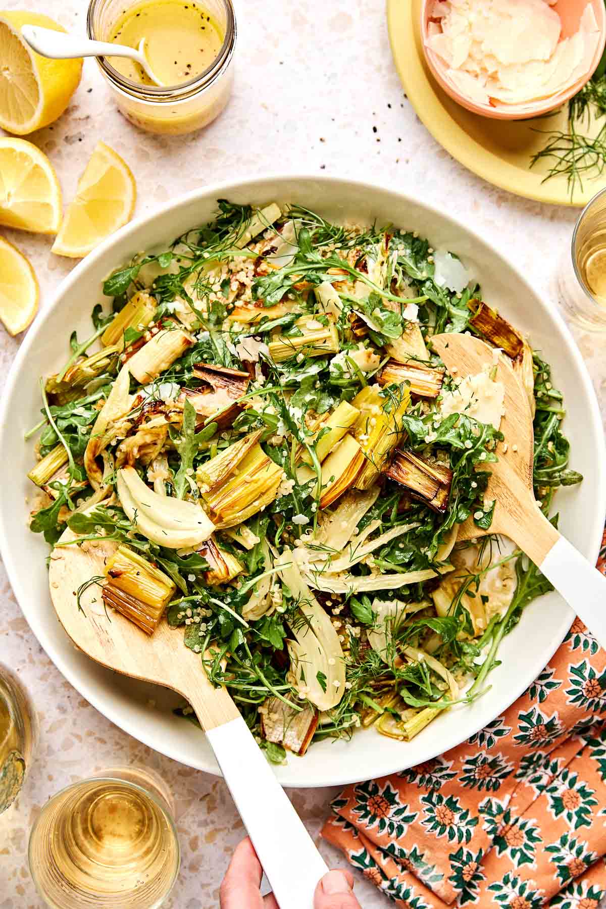 A bowl of roasted vegetable salad with arugula, quinoa, and herbs, served with wooden salad servers. Lemon wedges, dressing, and a floral napkin are nearby on a light table.