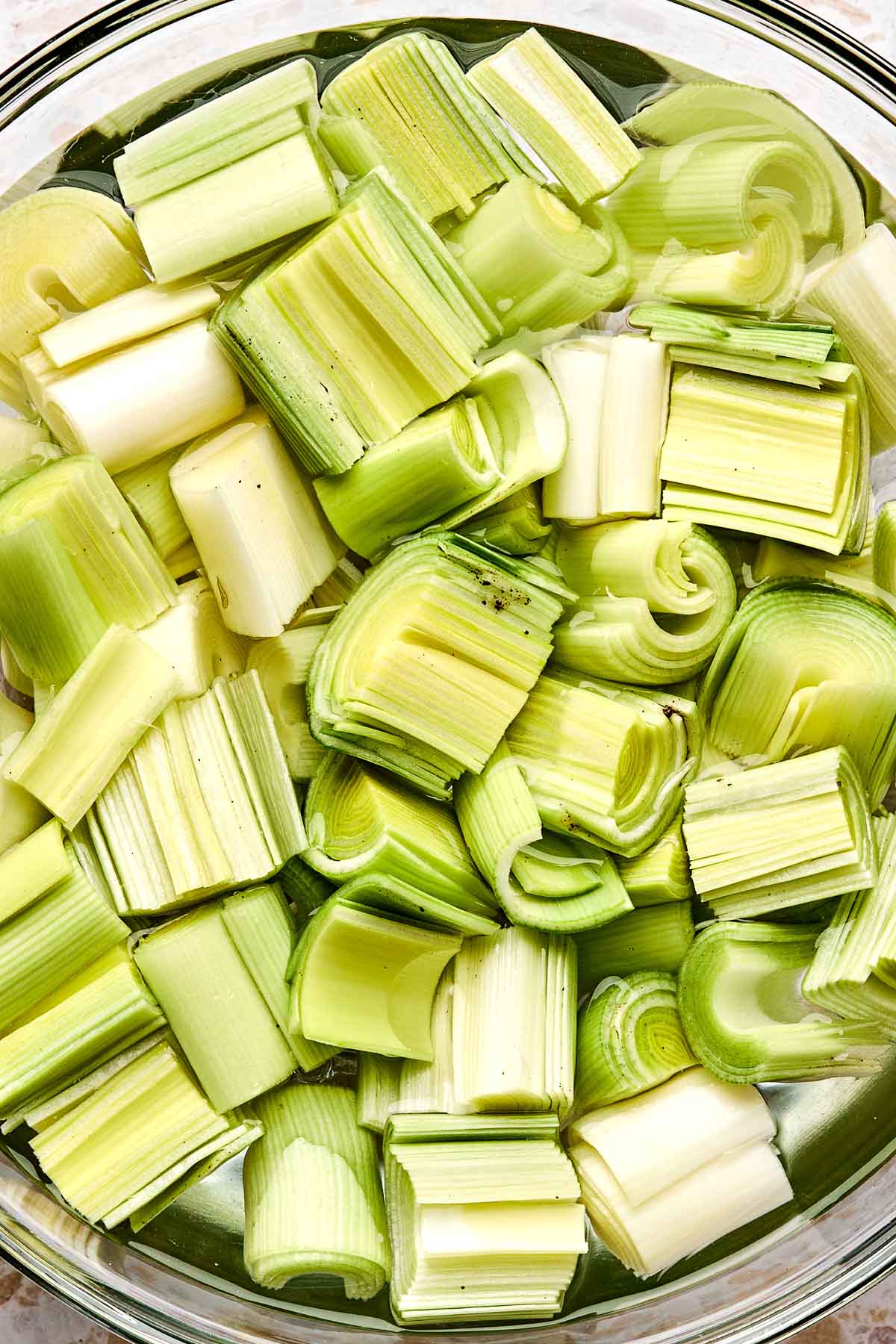 A close-up of chopped leeks soaking in a glass bowl of water, with light green and white pieces arranged in layers.