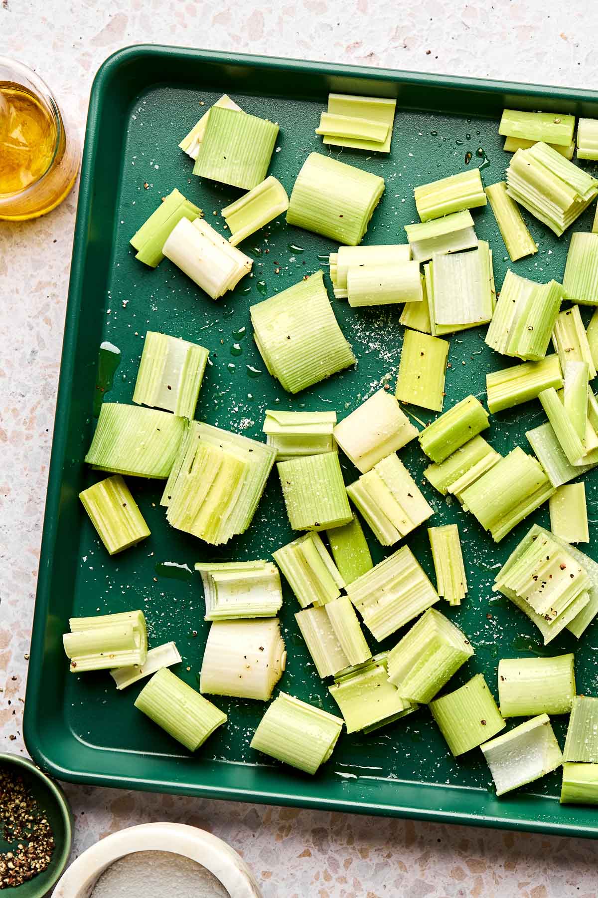 Chopped leeks spread out on a green baking tray, seasoned with salt, pepper, and oil, ready to be roasted. Small bowls of oil, salt, and pepper are beside the tray on a light countertop.
