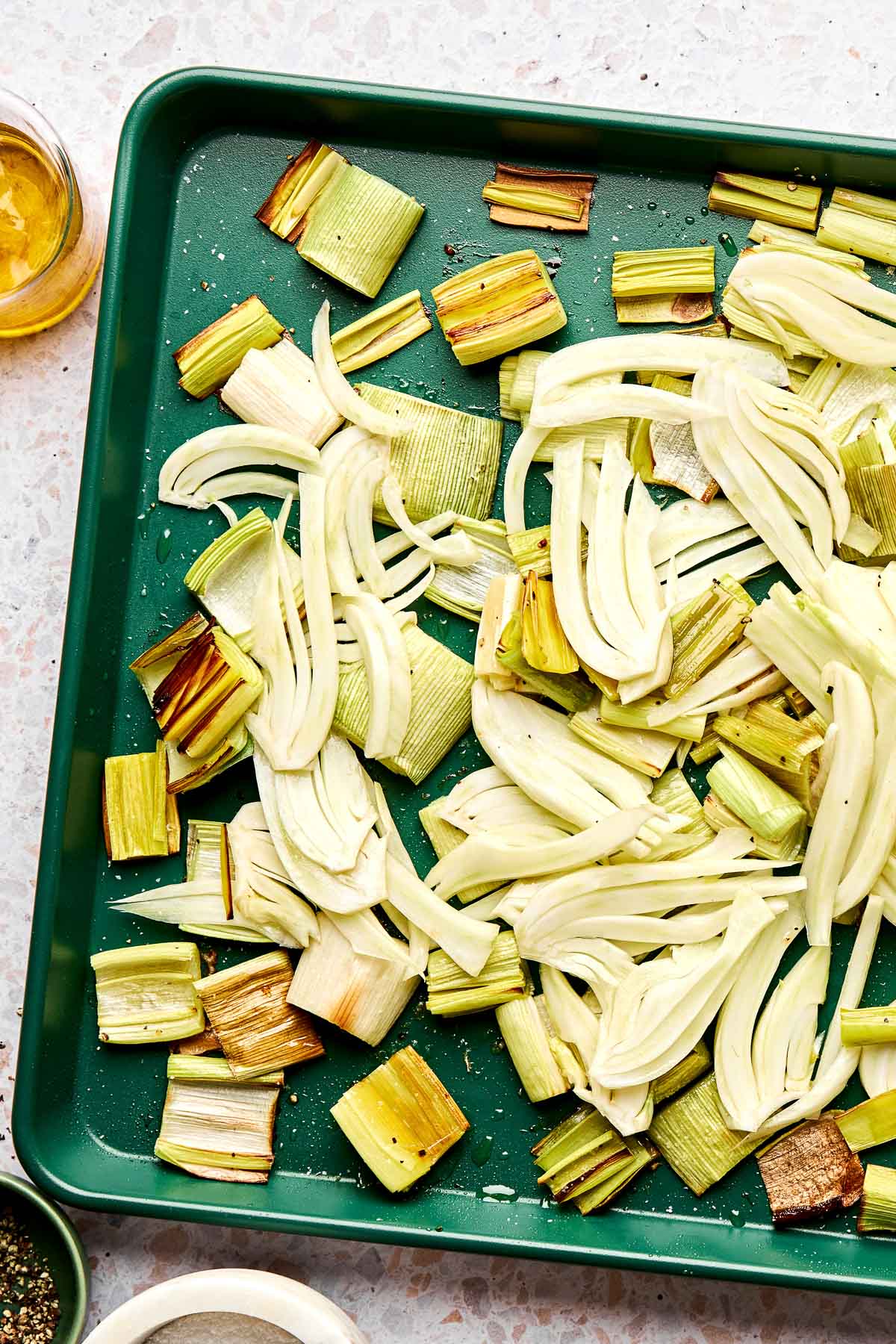 A green baking tray with sliced fennel and leeks, lightly seasoned, ready to be roasted. The tray sits on a light countertop with small bowls of oil and seasonings nearby.
