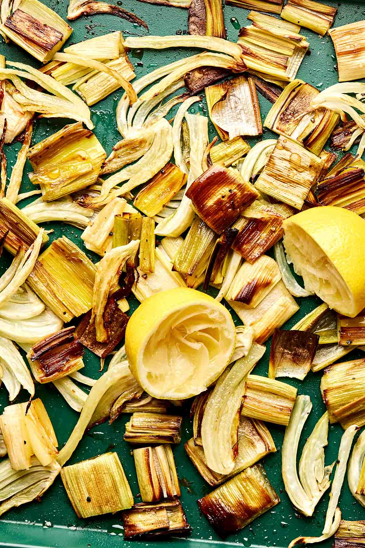 Roasted chopped leeks, fennel slices, and two squeezed lemon halves spread on a green baking tray.