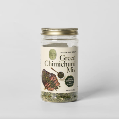 A clear glass jar with a gold lid labeled Green Chimichurri Mix. The label shows grilled meat topped with chimichurri and reads, “Just add oil & vinegar in minutes.” Some mix is visible at the bottom of the jar.