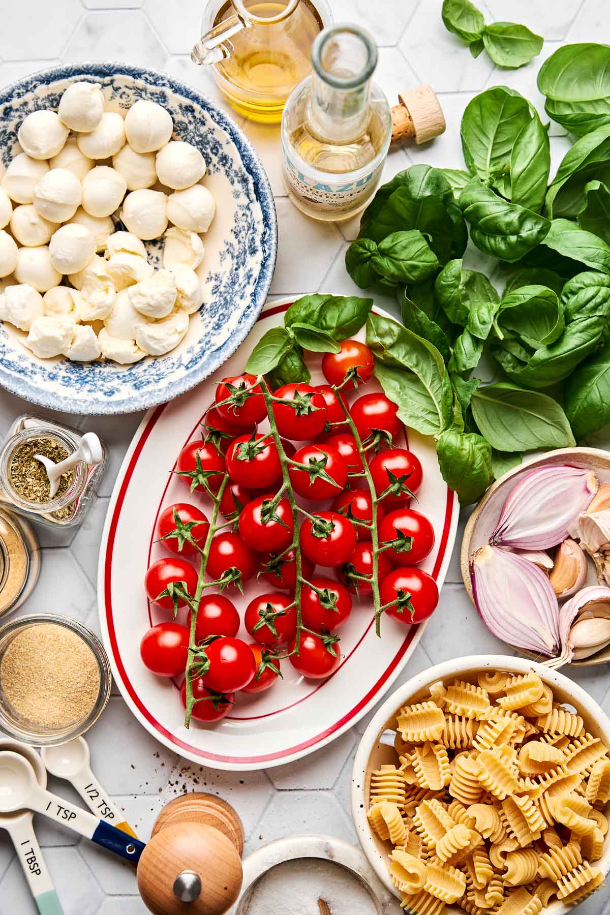 A flat lay of fresh ingredients including vine tomatoes, basil leaves, mozzarella balls, dry pasta, garlic, shallot, spices, olive oil, and measuring spoons arranged on a white surface.