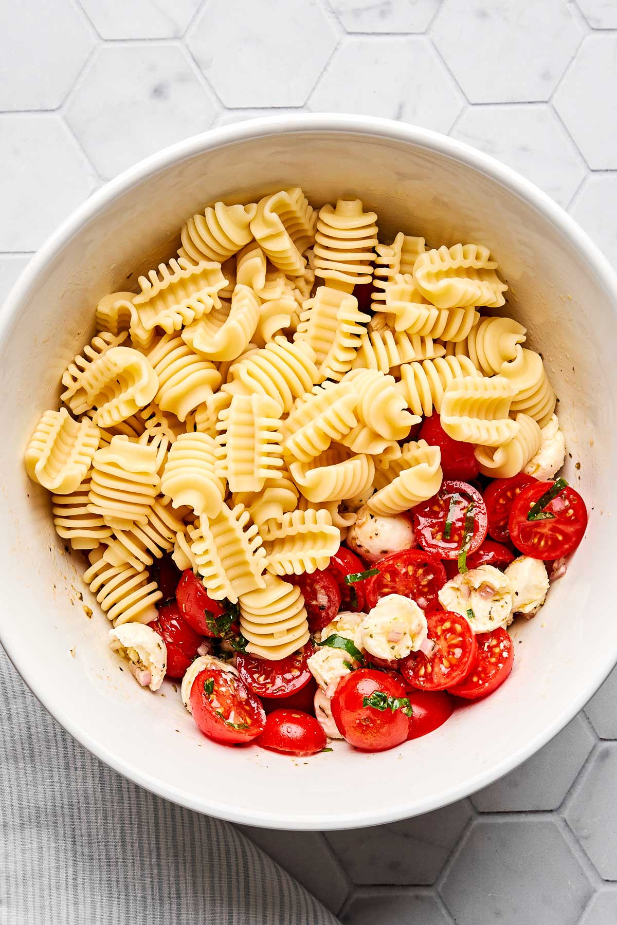 A white bowl containing uncooked radiatore pasta, halved cherry tomatoes, mozzarella balls, and chopped herbs on a light-colored hexagon tile surface.