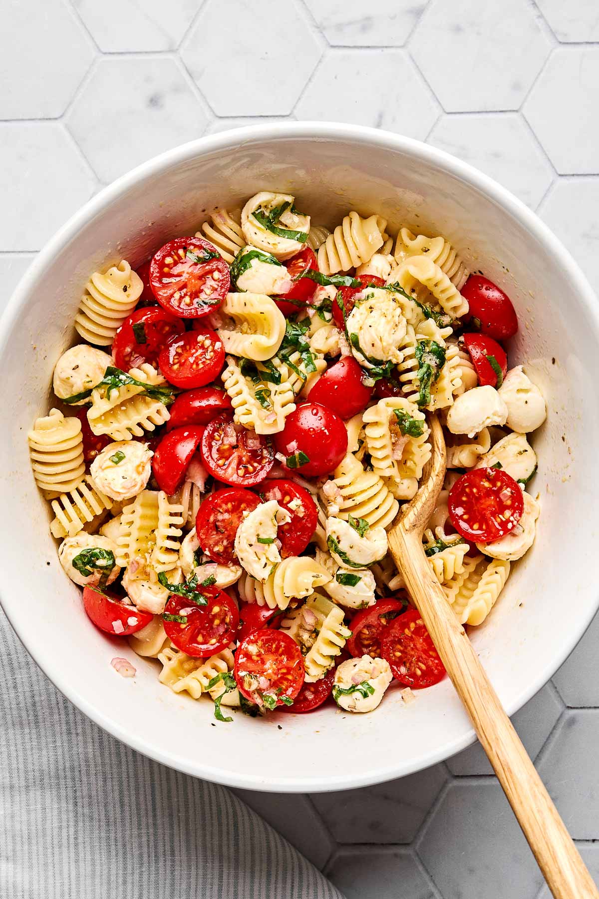 A white bowl filled with short pasta, halved cherry tomatoes, mozzarella balls, chopped basil, and a wooden spoon on a light hexagon-tiled surface.