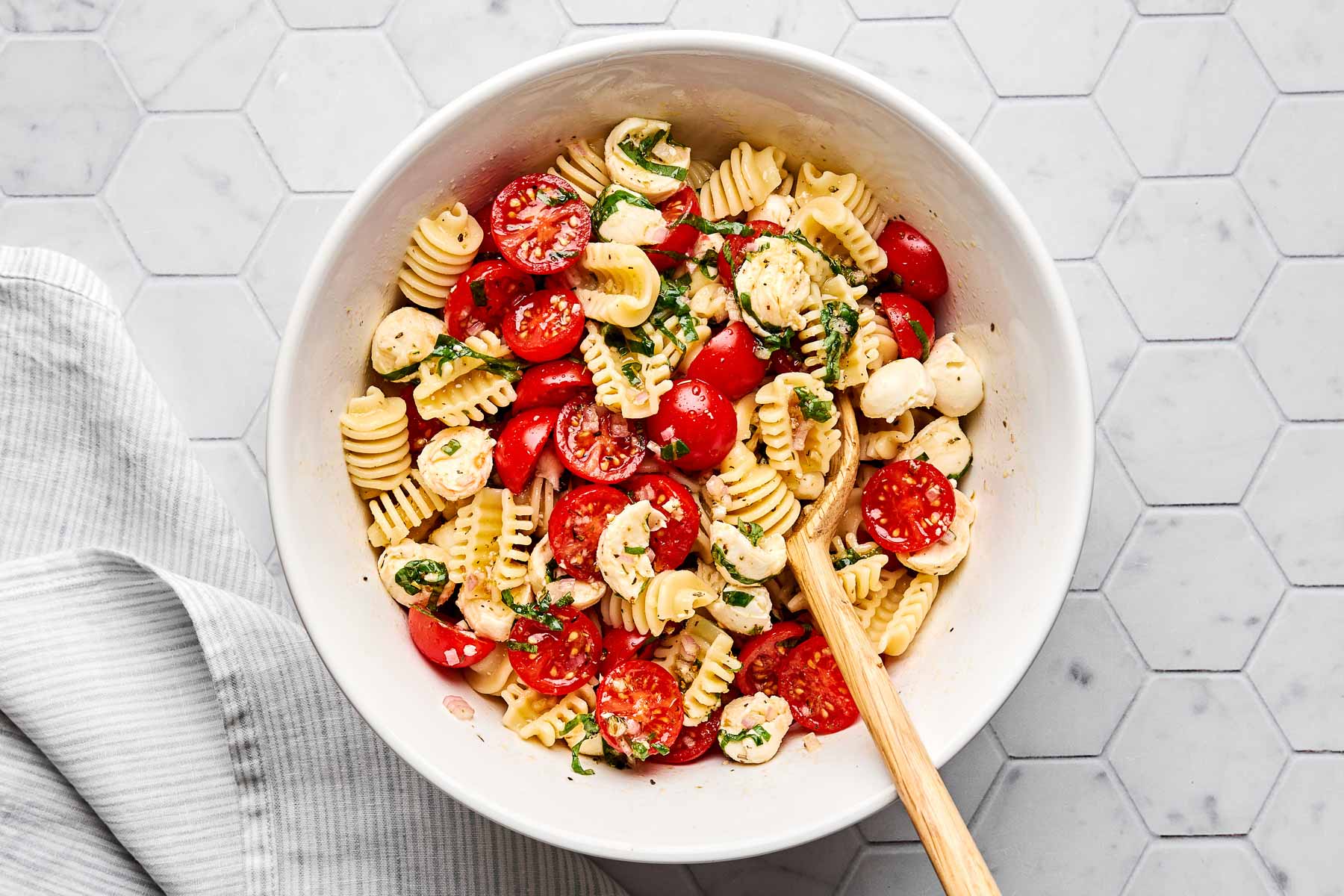 A white bowl filled with pasta salad, featuring spiral pasta, cherry tomatoes, mozzarella balls, and fresh basil, sits on a light hexagon-tiled surface with a wooden spoon and a striped cloth nearby.
