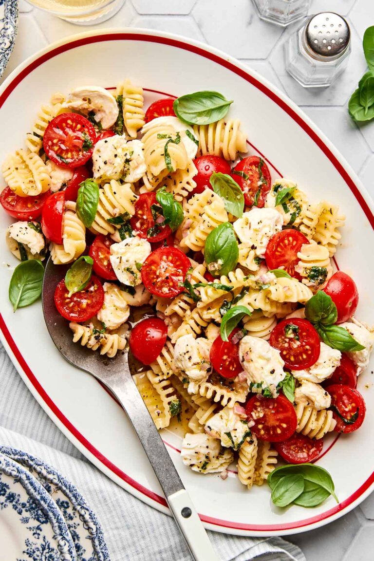 A serving platter with radiatore pasta, cherry tomatoes, mozzarella chunks, and fresh basil leaves. A large serving spoon rests on the dish. The background includes a salt shaker and a sprig of basil.