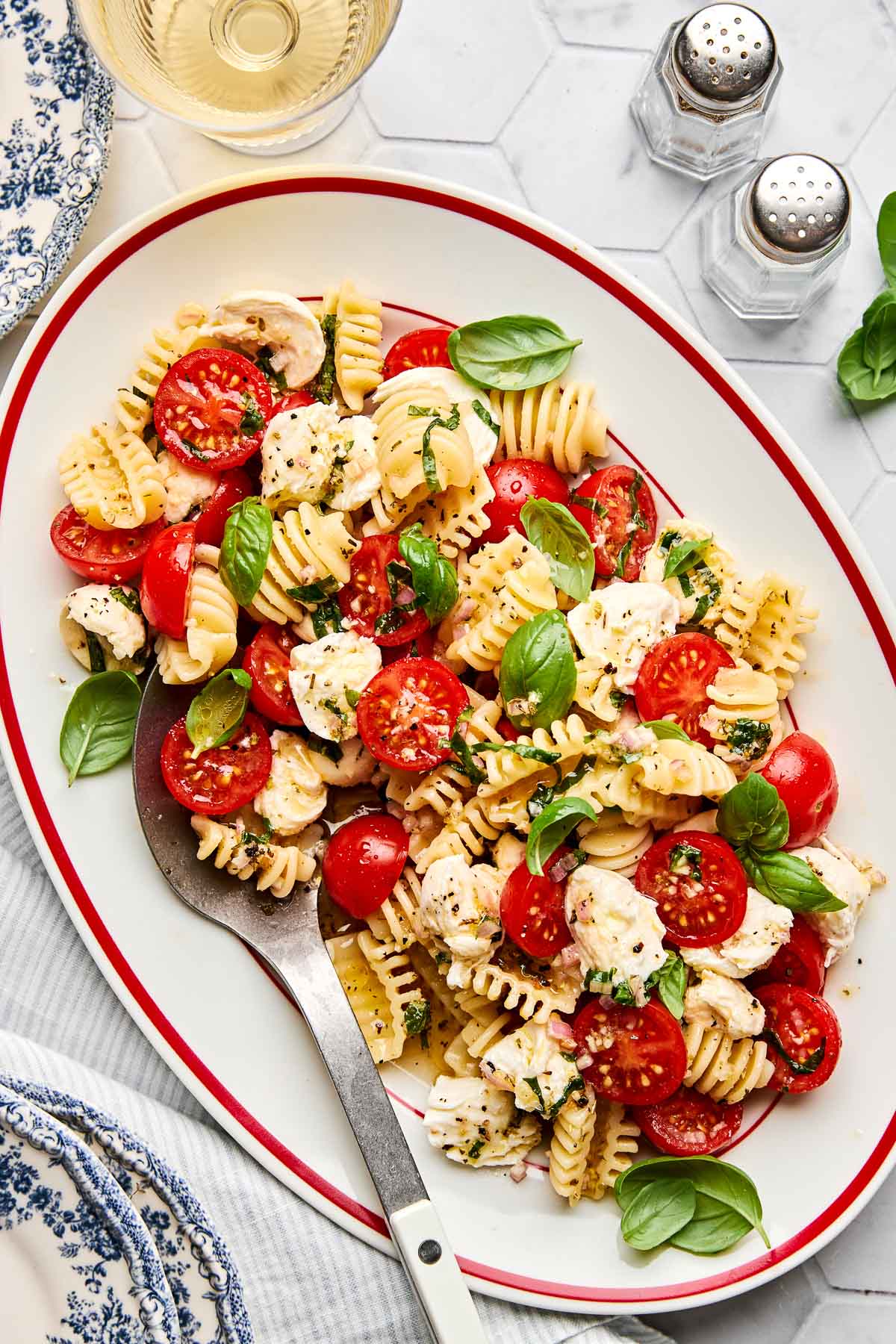 A platter of pasta salad with short noodles, cherry tomatoes, mozzarella balls, and fresh basil leaves, garnished with herbs. A large serving spoon rests on the plate; salt and pepper shakers are visible nearby.