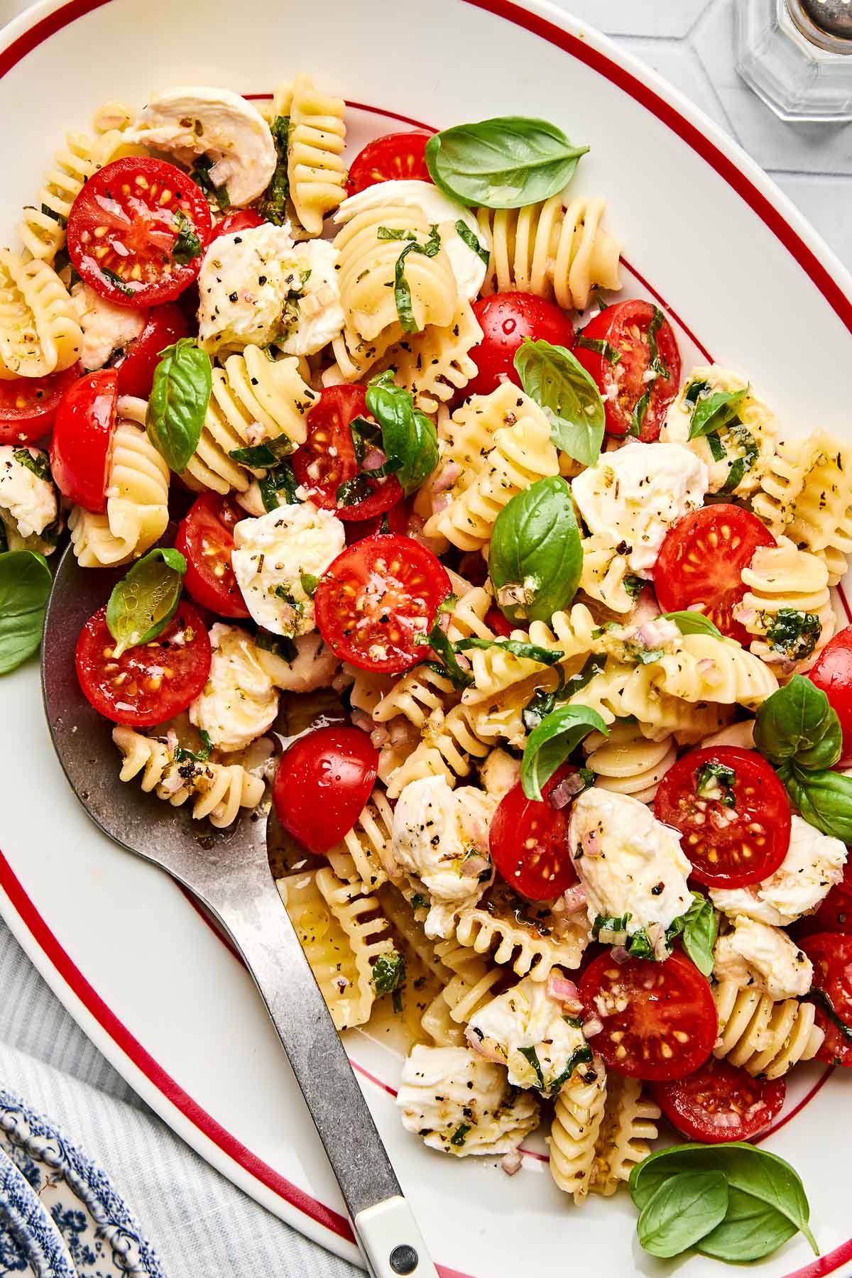 A platter of pasta salad with radiatore, cherry tomatoes, mozzarella balls, fresh basil leaves, and a sprinkle of herbs, served with a large spoon.