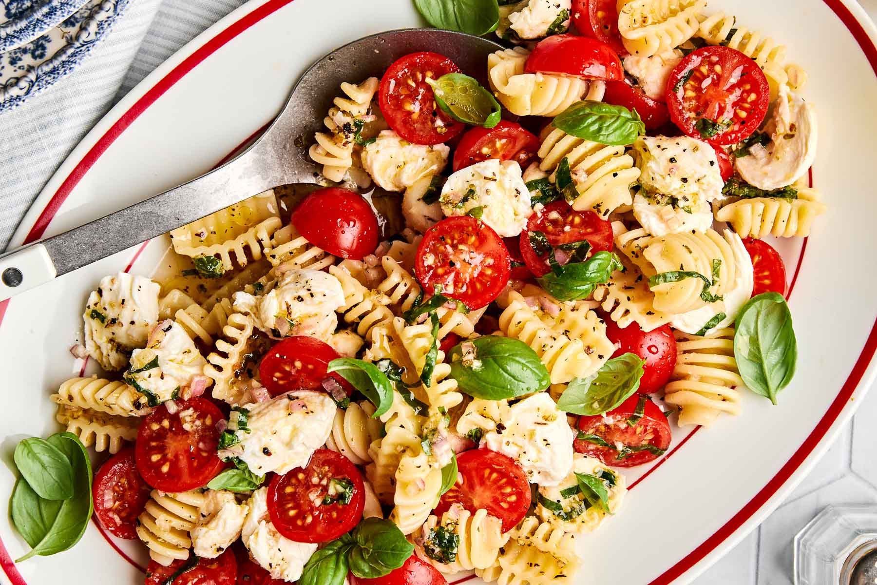 A plate of pasta salad with rotini noodles, halved cherry tomatoes, mozzarella pieces, fresh basil leaves, and a sprinkle of herbs, served with a large metal spoon on the side.