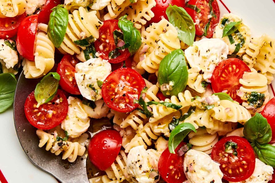 A close-up of a pasta salad with radiatore, cherry tomatoes, mozzarella balls, fresh basil leaves, and a light herb dressing on a white plate.