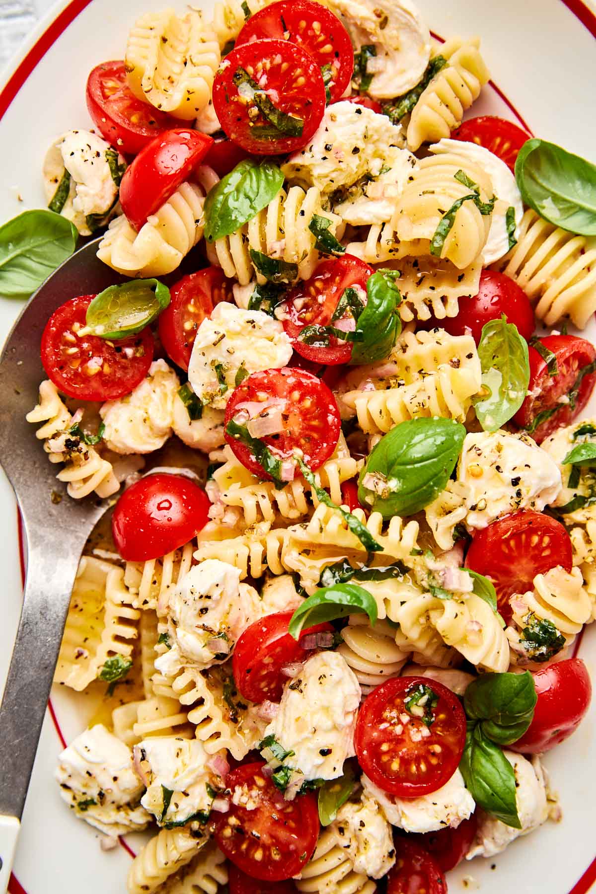 Pasta salad with radiatore pasta, halved cherry tomatoes, mozzarella chunks, fresh basil leaves, and a sprinkle of black pepper, served on a white plate with a spoon.