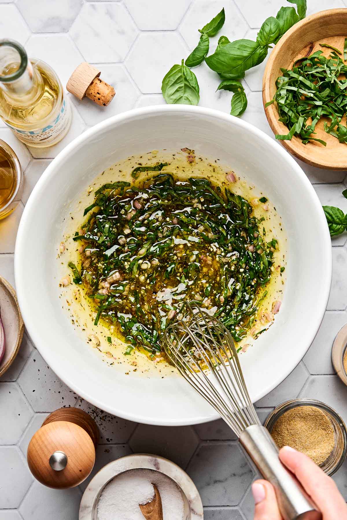 A hand whisks a mixture of olive oil, chopped herbs, and shallots in a white bowl surrounded by fresh basil, chopped herbs, seasonings, and a bottle of olive oil on a light countertop.