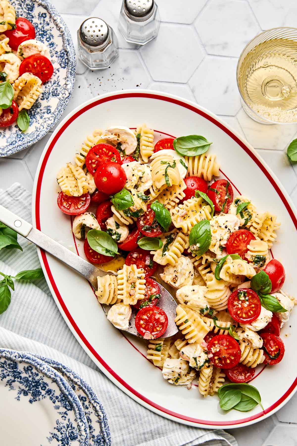 A platter of pasta salad with cherry tomatoes, mozzarella, basil, and black pepper sits on a white surface. There are two blue-patterned plates, salt and pepper shakers, and a glass of white wine nearby.