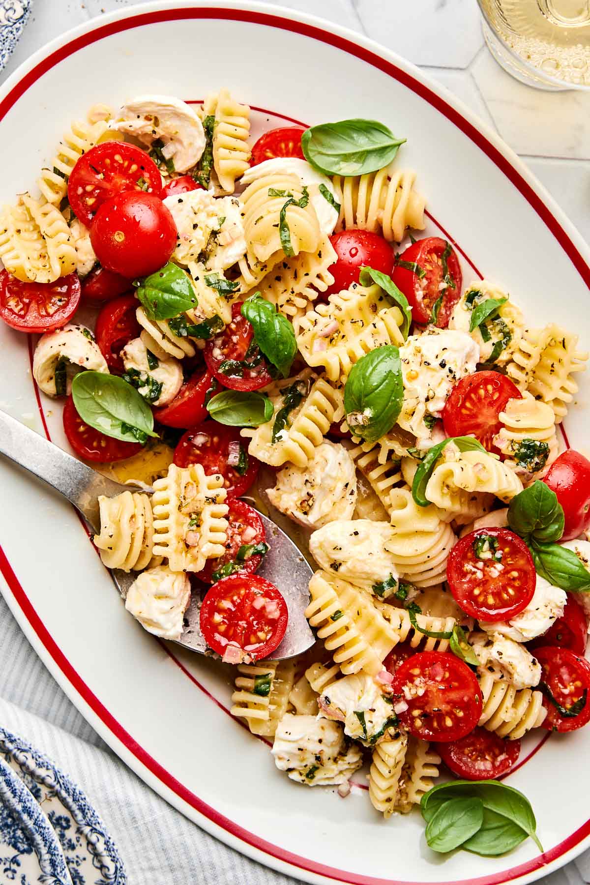A platter of pasta salad with short noodles, halved cherry tomatoes, mozzarella balls, fresh basil leaves, and a sprinkle of black pepper, served with a large serving spoon.