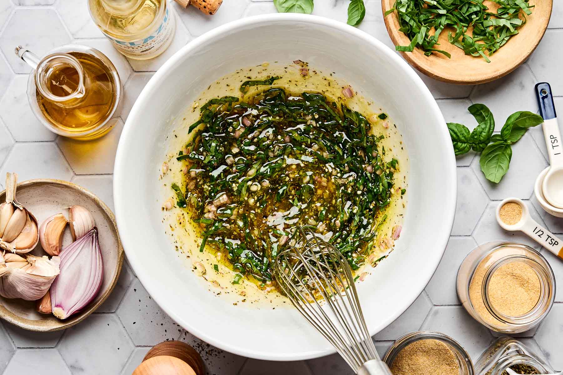 A bowl of homemade vinaigrette being whisked, surrounded by fresh basil, garlic, shallot, olive oil, spices, and measuring spoons on a white hexagonal tile countertop.