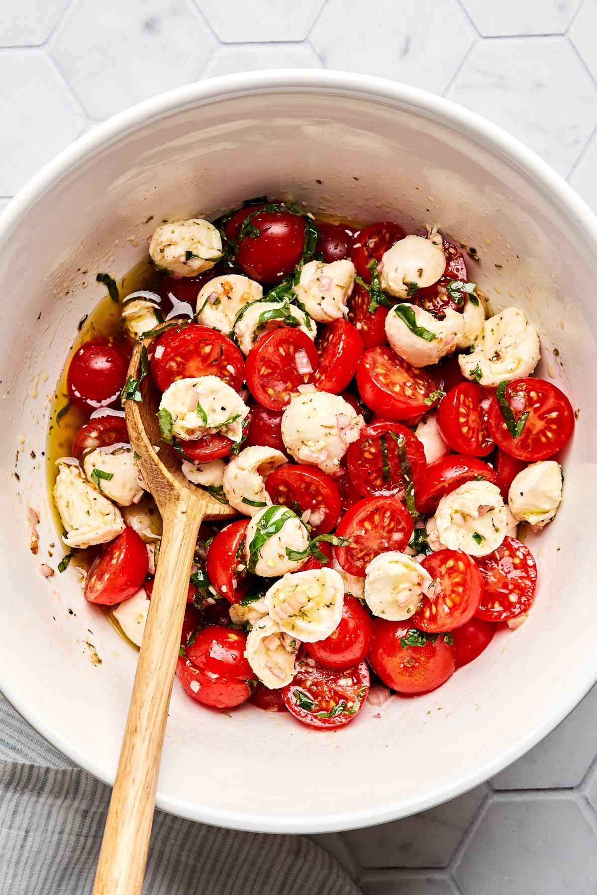 A white bowl filled with a salad of halved cherry tomatoes, mozzarella balls, chopped basil, and dressing, with a wooden spoon resting inside. The bowl sits on a light-colored, hexagonal tile surface.