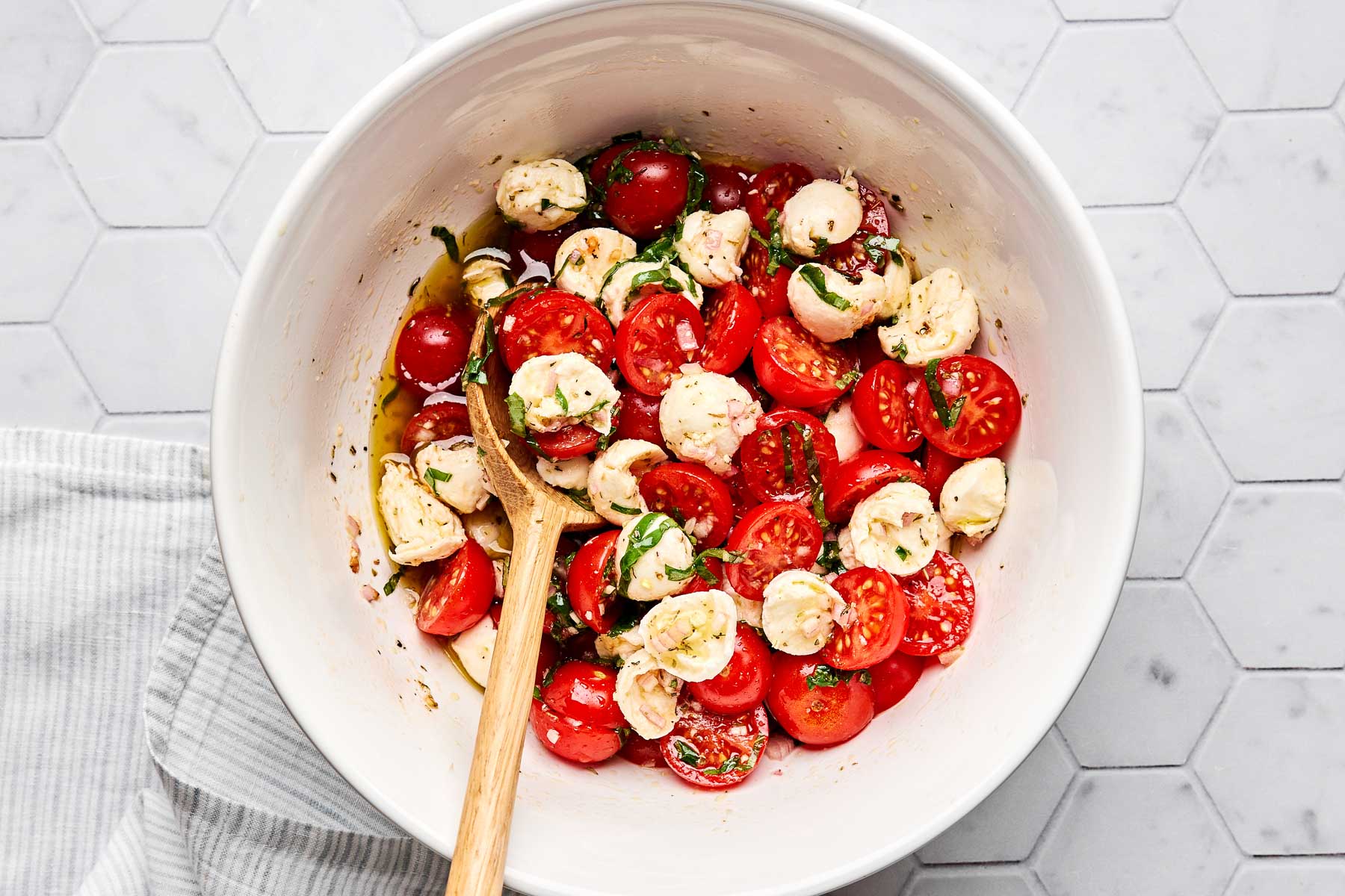 A white bowl filled with halved cherry tomatoes, mozzarella balls, and fresh basil, mixed with olive oil, sits on a hexagon-tiled surface. A wooden spoon rests inside the bowl.