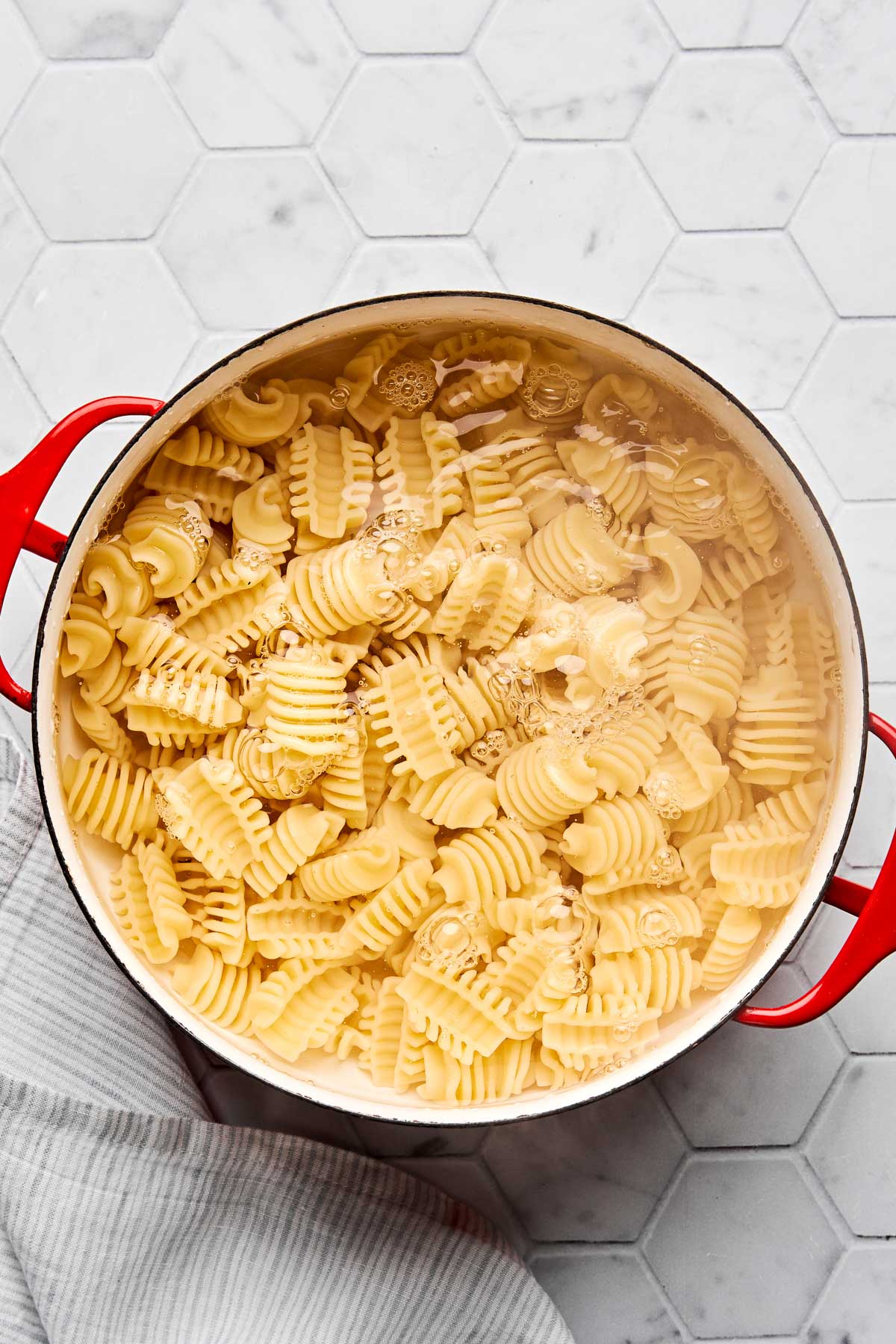 A red pot filled with boiling water and radiatore pasta sits on a white hexagonal-tiled countertop, with a striped dish towel partially visible beside it.